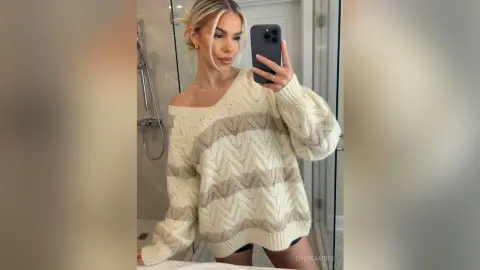 Blonde woman in an oversized cream cable-knit sweater taking a mirror selfie in a bright bathroom.