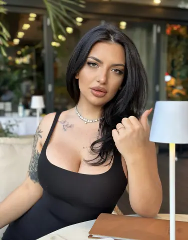 NoraSin wearing a black tank top and silver necklace, posing with her hand near her face while sitting at a table outdoors.
