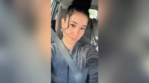 JuliahLewis poses in a car wearing a grey striped long-sleeve top with her hair in a high bun.