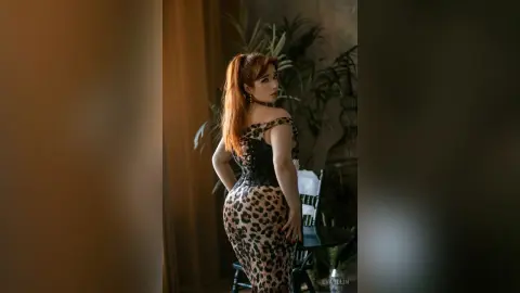 Elegant redhead woman in a leopard print dress posing in a moody, dimly lit indoor setting.