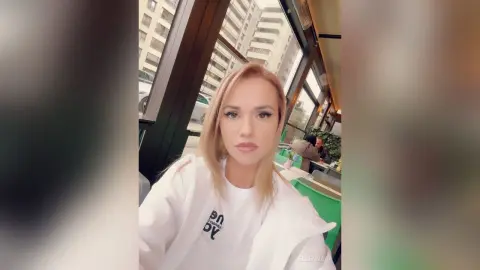 Blonde woman in a white sweatshirt sitting by a bus window with a city view and soft natural lighting.
