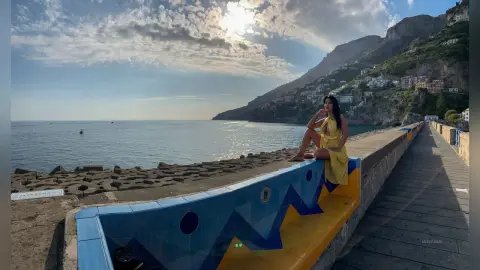 MissyJolie sits on a blue and yellow tiled wall overlooking the ocean, wearing a short yellow sundress.