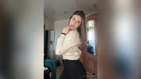 Young woman in a cream sweater and black pants posing thoughtfully in a bright modern living room.