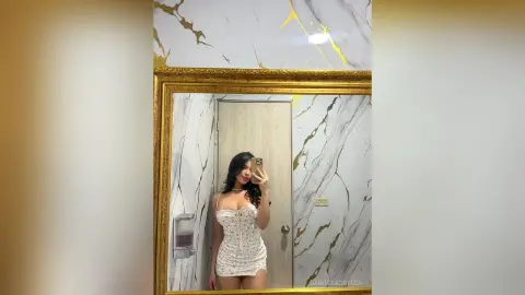 GabrielaOrtega poses in a mirror wearing a white floral print bodysuit, showing off her long dark hair and cleavage against a marble wall background.
