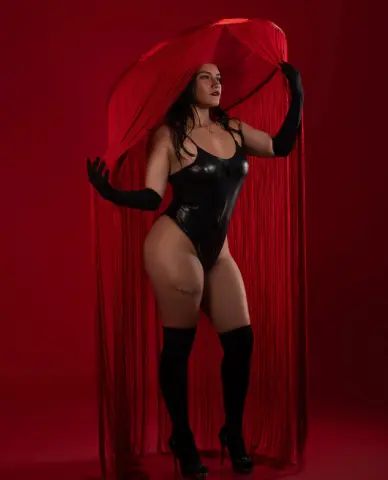 Curvy woman in a black bodysuit and gloves posing against a dramatic red fabric backdrop.