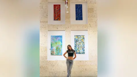 KateGale stands in front of large colorful paintings, wearing a black crop top and light blue jeans with her hands on her hips.