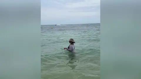 Woman in a sun hat wading through calm turquoise ocean water on a peaceful summer day.