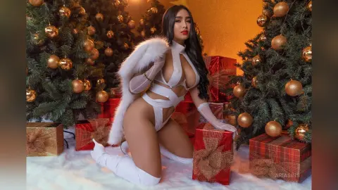 Woman in white angel costume and wings kneeling among Christmas trees and red gift boxes in a festive mood.