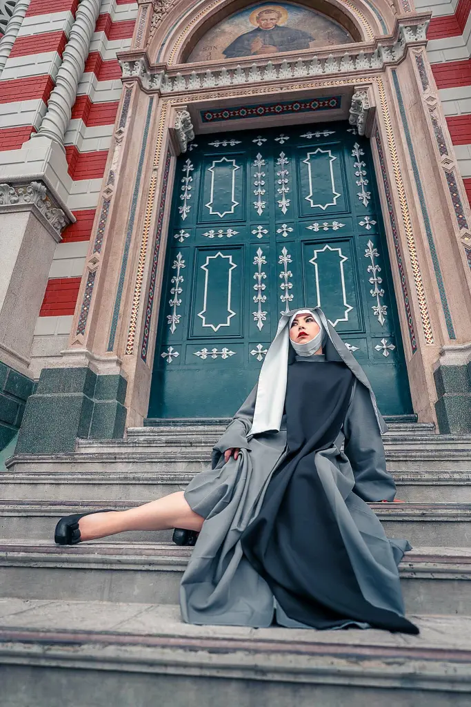 NikolleWilliams — Woman in a grey and black nun habit sitting on stone steps of an ornate cathedral entrance.