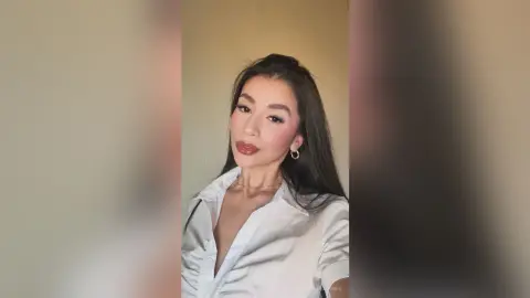 BellaaJones poses in a white button-down shirt with her long dark hair down and gold hoop earrings.