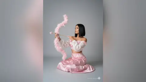 Elegant woman in a pink satin outfit holding a whimsical pink feathered bow against a grey studio background.