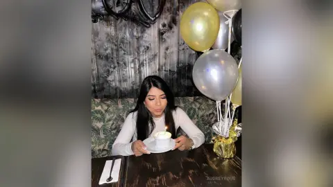 Young woman in a white long-sleeve shirt celebrating a birthday with a lit candle and silver balloons.