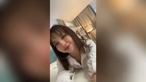 Smiling young woman in a white button-down shirt relaxing in a cozy, sunlit bedroom.