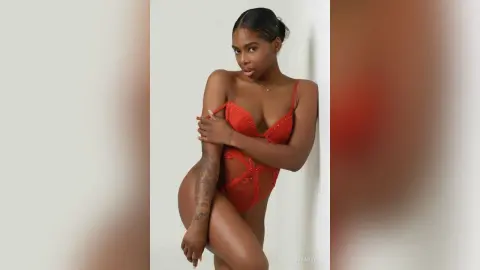 Confident Black woman in a red lace bodysuit posing against a minimalist white wall background.
