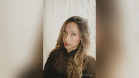 LaurentBelluci looks at the camera with long wavy hair and a black sheer blouse against a white curtain background.