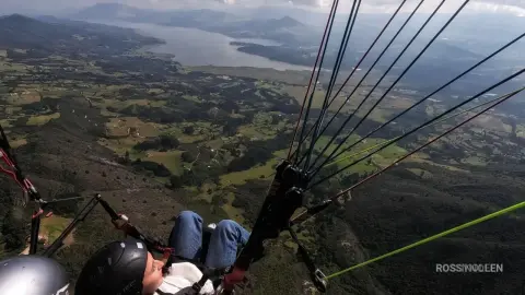 RossiNoleen paraglides high above a lush green valley and a large lake, wearing a white top and blue jeans.