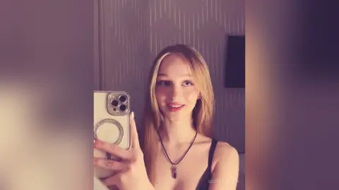 Young woman with long blonde hair wearing a black tank top taking a mirror selfie in a modern room.
