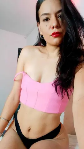 Young woman in a pink crop top and black bikini bottoms sitting in a bright, minimalist room.