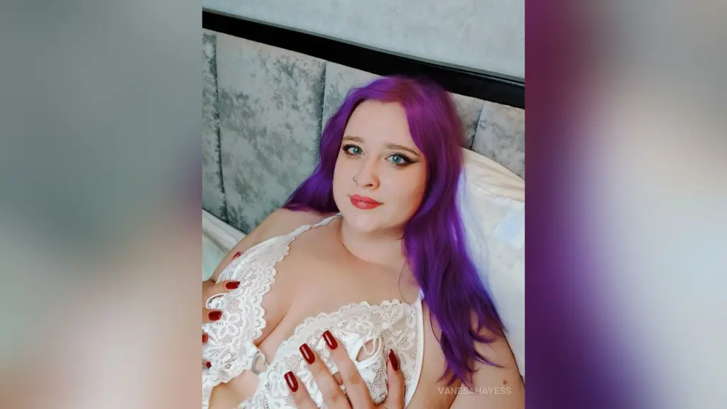 Confident plus-size woman with purple hair wearing black lingerie and fishnet tights in a studio setting.