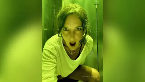 MichaelaBelle looks directly into the camera with an open mouth, wearing a white t-shirt and a silver necklace against a green background.
