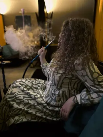 Curly-haired woman in a patterned maxi dress smoking a hookah in a cozy, dimly lit room.