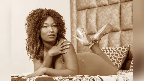 Curvy woman in a bodysuit relaxing on a bed with high heels in a warm, cozy bedroom setting.