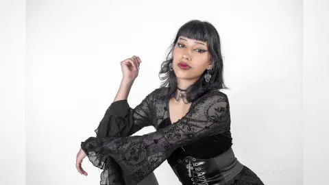 LilithBrooks poses in a black lace long-sleeve top and black corset against a white background.