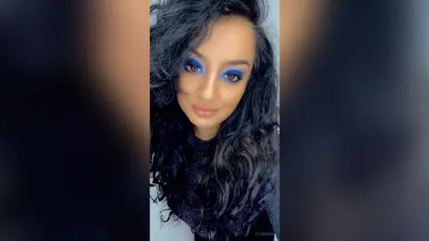 Woman with long wavy dark hair and bold blue eyeshadow wearing a black top in a soft indoor setting.