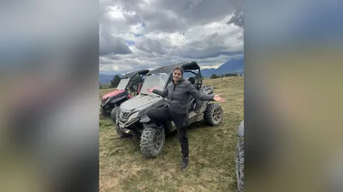 Happy woman in a black jacket standing by an off-road ATV in a scenic mountain landscape under a cloudy sky.