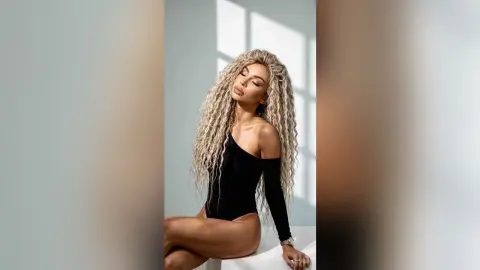 Elegant woman with long blonde curly hair wearing a black bodysuit in a bright, minimalist studio setting.