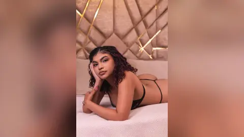 Young woman in black lace lingerie relaxing on a white bed in a luxurious, warm-lit bedroom setting.