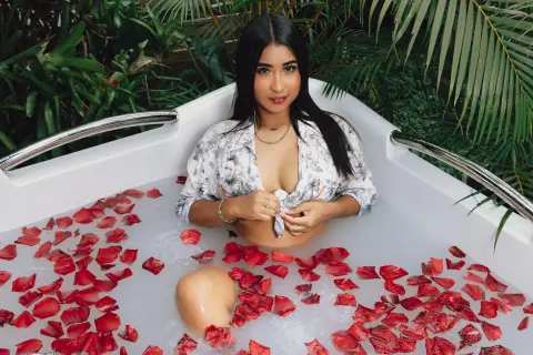 Young woman in a white floral shirt relaxing in a luxury bathtub filled with red rose petals in a tropical garden.