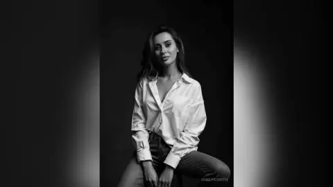Elegant woman in a white button-down shirt posing in a moody black and white studio portrait.
