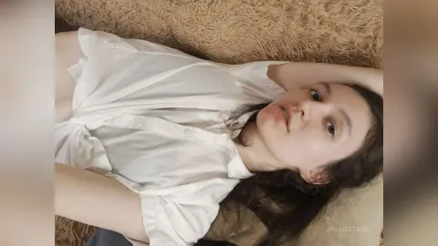 MillieStrife lies on a tan shag rug wearing a white button-up shirt, looking at the camera with her dark hair spread out.