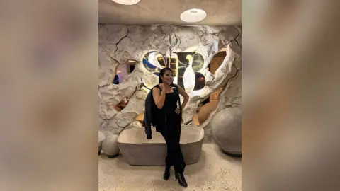 Stylish woman in an all-black outfit posing confidently in a modern, sculptural luxury lounge setting.
