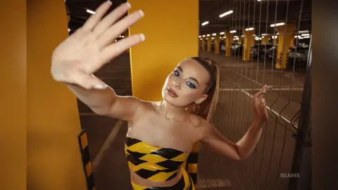BeaNix poses in a yellow parking garage wearing a black and yellow striped tube top, reaching a hand toward the camera.