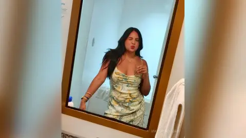 Smiling woman in a floral sundress posing in a bathroom mirror for a casual lifestyle portrait.