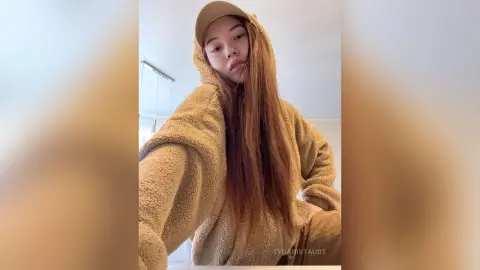 Young woman in a cozy tan fleece hoodie and baseball cap relaxing in a bright indoor setting.