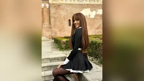 TiffanyAddams smiles while sitting outdoors in a black and white schoolgirl outfit with long brown hair and black tights.