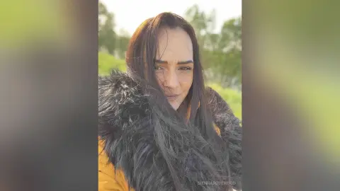 Smiling woman in a warm mustard yellow coat and black faux fur collar outdoors in a sunny park setting.