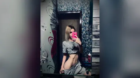 Fit woman in a grey crop top and sweatpants taking a mirror selfie in a modern bathroom.