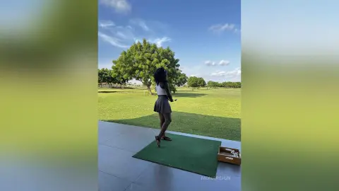 Young woman in a black tank top and skirt standing on a patio overlooking a lush green field under a blue sky.