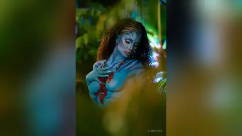 Blue-skinned Na'vi woman in tribal jewelry posing in a lush, mystical jungle setting.