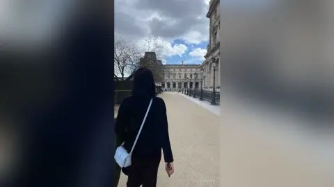 Woman in black outfit with white crossbody bag walking toward a grand historic palace under a cloudy sky.