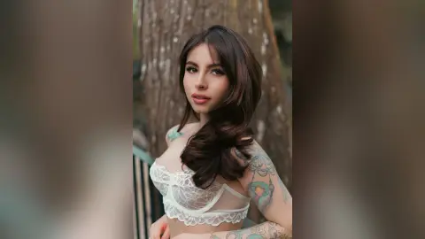 Tattooed woman in white lace lingerie posing outdoors in a soft, natural garden setting.