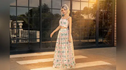 Blonde woman in a floral print crop top and maxi skirt standing on a terrace during a golden hour sunset.