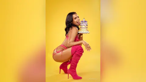 Playful woman in a pink bodysuit and tall boots holding a birthday cake against a bright yellow background.