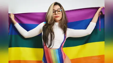 JessieLiu poses in a white long-sleeve top and a rainbow striped bodysuit while holding up a large pride flag behind her.