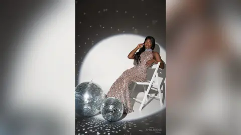 Glamorous woman in a sparkling sequin gown sitting by disco balls in a moody studio setting.