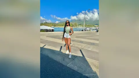 Stylish woman in a green bodysuit and white shorts standing on a sunny waterfront boardwalk under a blue sky.